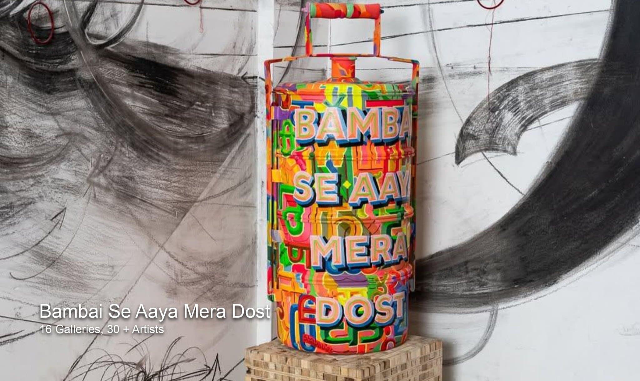 A brightly painted tiffin carrier lettered with the exhibition title, standing in front of a large charcoal wall drawing.