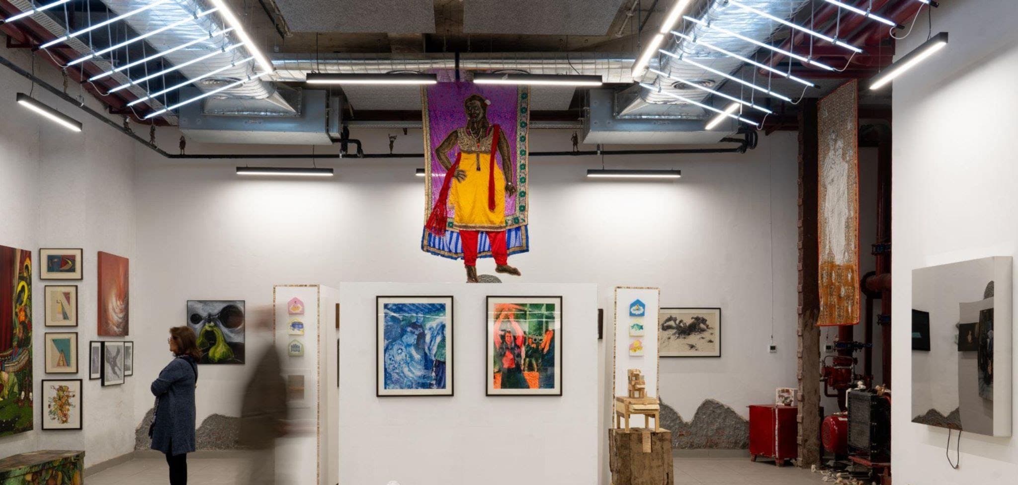 The gallery at Method Delhi with framed works on white walls and a textile figure suspended from the ceiling.